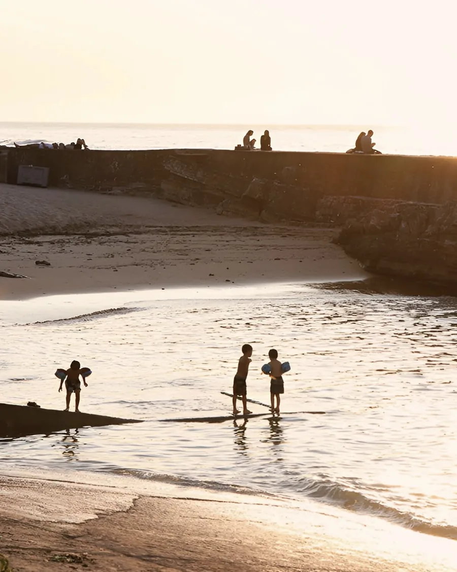Children in swim floaties paddling in the shallows at sunset, with people sitting along a stone breakwater behind