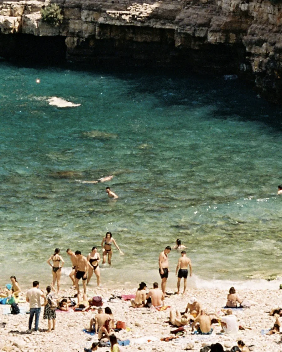 Swimmers and sunbathers on a pebble beach below limestone cliffs, clear turquoise water