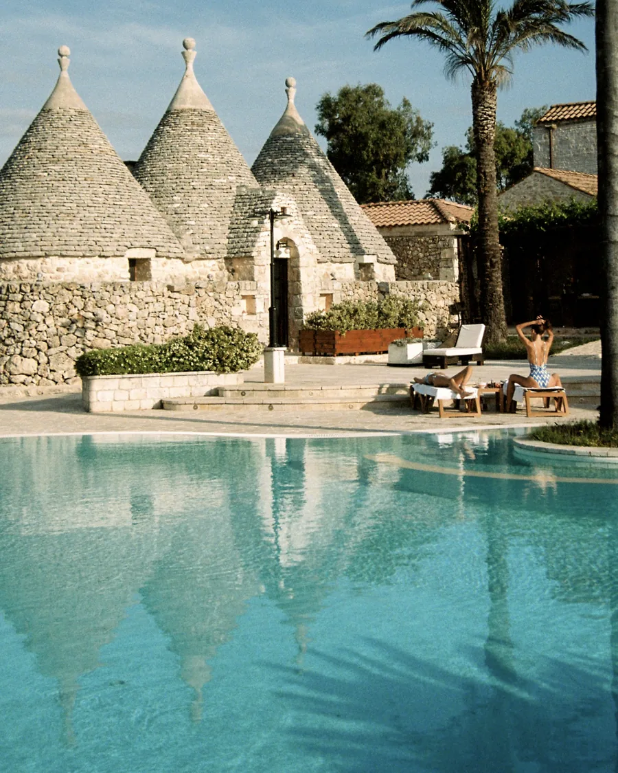 Two women on sun loungers beside a pool, stone trulli with conical roofs reflected in the water
